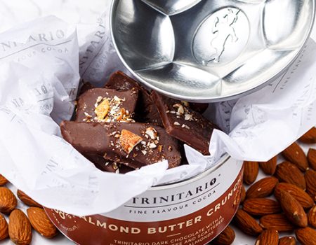 Pieces of almond butter crunch chocolate made with single-origin chocolate in a decorative tin, surrounded by whole almonds, with a silver lid resting nearby. | Trinitario