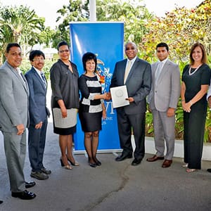 A group of seven people in business attire stand outdoors, posing with a certificate in front of a blue banner—celebrating excellence in hand-crafted chocolate made from fine flavour cocoa. | Trinitario