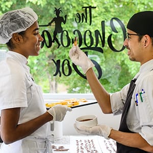 Two pastry chefs in uniform and gloves stand by a window; one feeds the other a treat made from fine flavour cocoa, with chocolates and tools arranged on the table nearby. | Trinitario