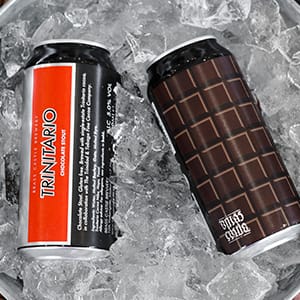 Two cans of Trinitario chocolate stout beer rest on ice; one with a red and silver label, the other with a brown design resembling chocolate squares—both brewed with sustainably grown, single-origin chocolate. | Trinitario