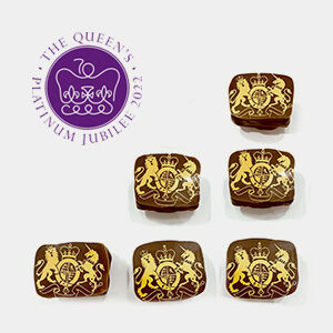 Six square handcrafted chocolates with gold royal crests are displayed below a purple emblem reading "The Queen's Platinum Jubilee 2022," featuring a crown and stylised text. Made with fine flavour cocoa for a luxurious treat. | Trinitario