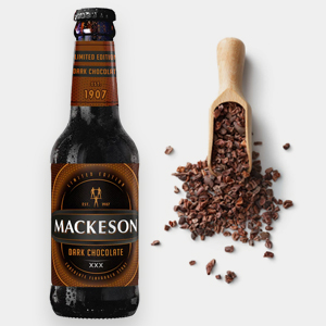 A bottle of Mackeson Dark Chocolate beer stands next to a wooden scoop filled with Trinitario chocolate cocoa nibs, some scattered on a white surface. | Trinitario