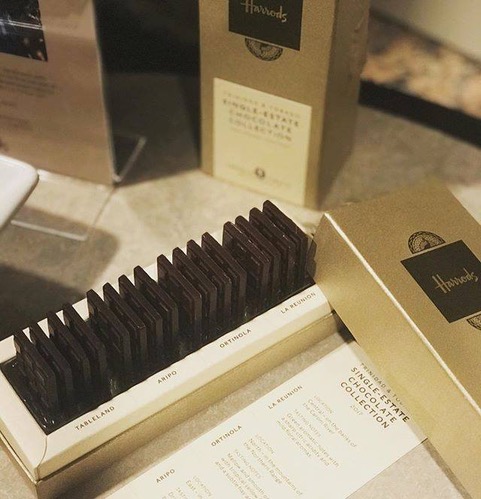 A gold Harrods box containing neatly arranged rectangular pieces of dark chocolate, with packaging and a label listing chocolate varieties displayed nearby. | Trinitario