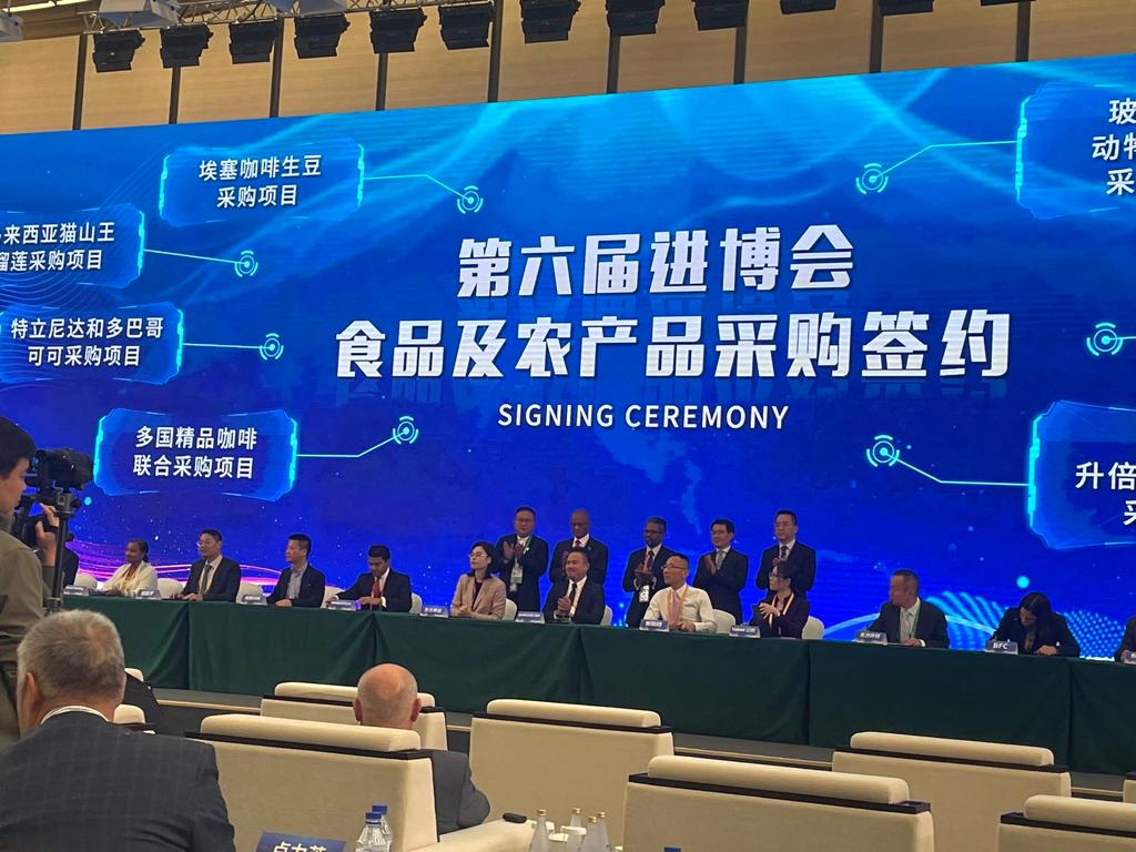A group of people sit at a long table on stage during a signing ceremony, with a large blue screen behind them displaying Chinese and English text, highlighting partnerships for sustainably grown chocolate and Caribbean cocoa. | Trinitario