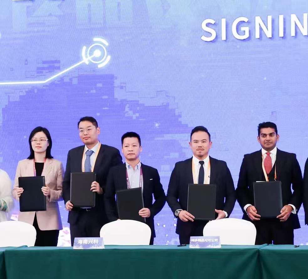 Five people in formal attire stand behind a table holding black folders, with a signing ceremony backdrop highlighting sustainably grown chocolate partnerships. | Trinitario
