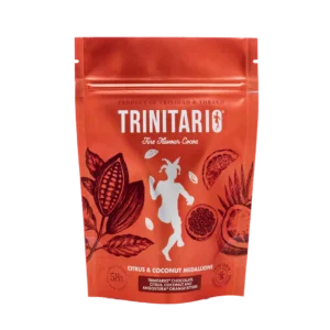 A red resealable pouch labelled "Trinitario Fine Flavour Cocoa" features cocoa pods, citrus fruits, and a dancing goat. The text highlights "Citrus & Coconut Medallions," handcrafted chocolate made with fine flavour cocoa. | Trinitario
