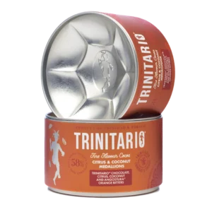 A round tin of single-origin chocolate Trinitario citrus and coconut medallions is shown, with the lid leaning against the container to display the logo and product details, highlighting its fine flavour cocoa origins. | Trinitario