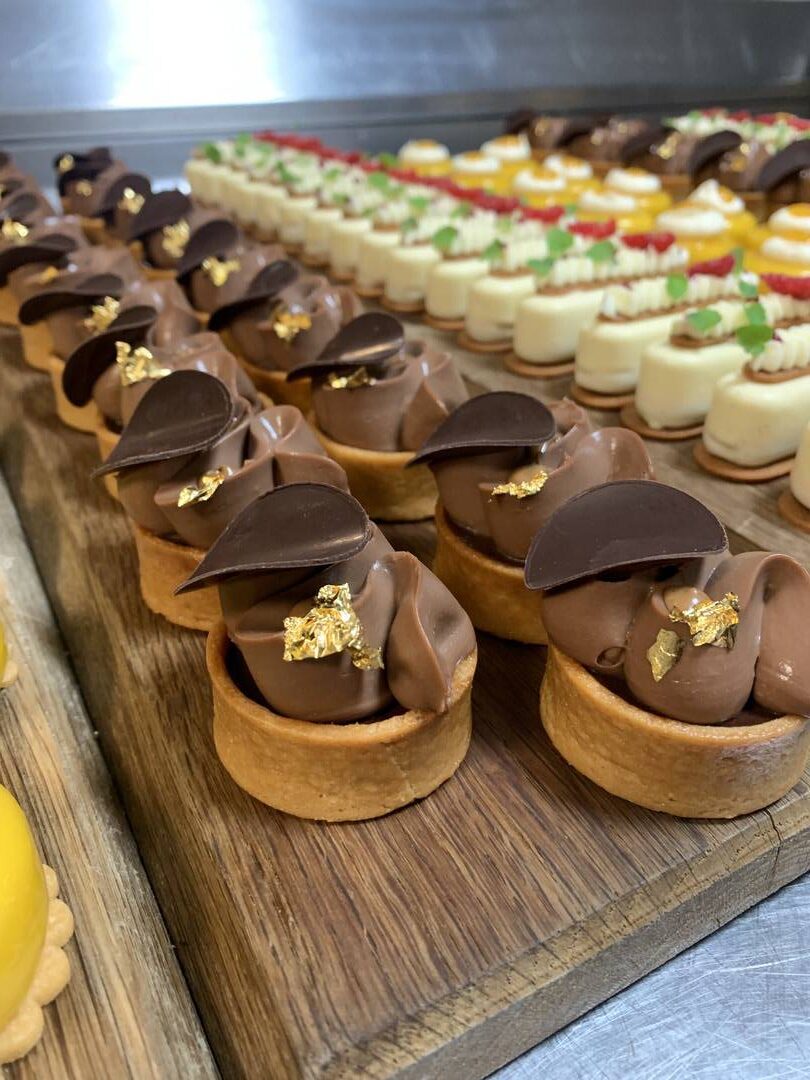 Rows of assorted mini desserts on a wooden board, featuring chocolate tarts made with fine flavour cocoa and gold leaf, alongside various colourful pastries in the background. | Trinitario