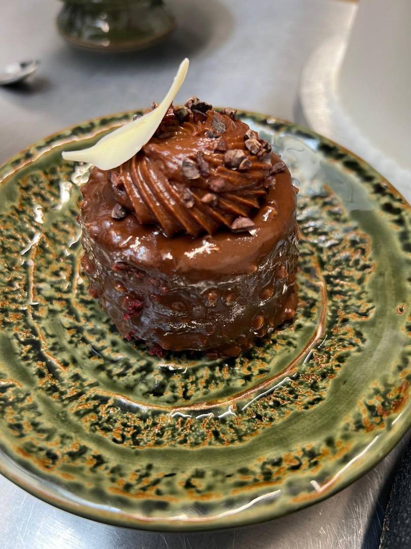 A small chocolate dessert made with single-origin chocolate, topped with icing, chocolate shavings, and a white chocolate piece, served on a green textured ceramic plate. | Trinitario
