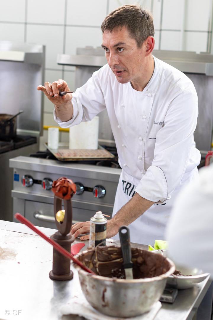 A chef in a white uniform demonstrates a chocolate technique using fine flavour cocoa in a kitchen, with a metal bowl of melted chocolate and a chocolate sculpture on the counter. | Trinitario