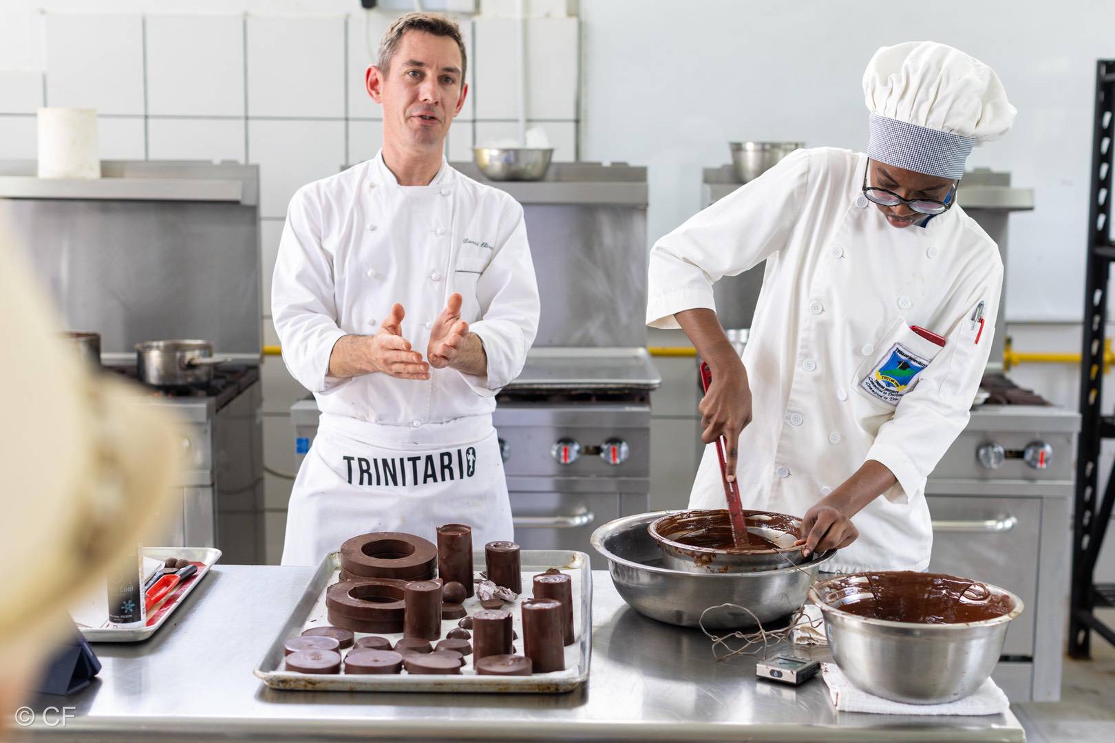 Two pastry chefs in a kitchen; one explains while the other stirs melted Trinitario chocolate in a bowl surrounded by chocolate moulds and kitchen equipment. | Trinitario