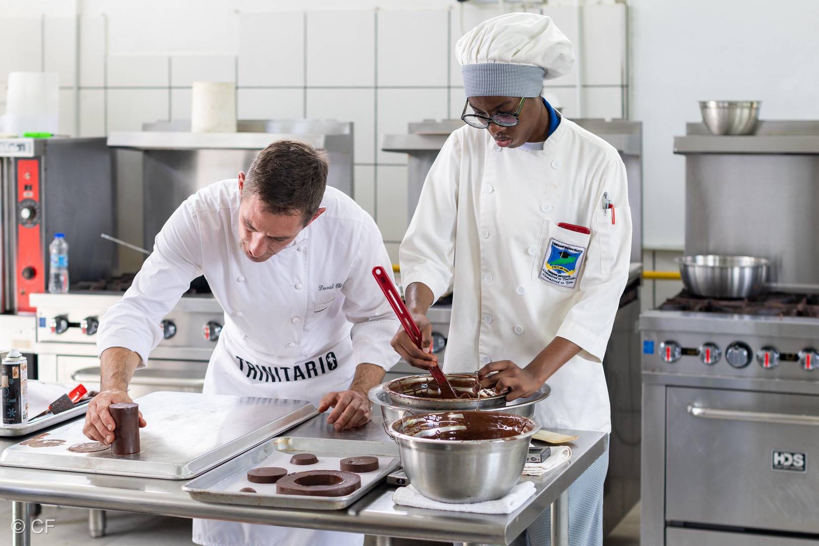Two chefs in a professional kitchen craft handmade chocolate; one pours chocolate into moulds while the other smooths it on a tray. Stainless steel bowls and utensils are on the table. | Trinitario
