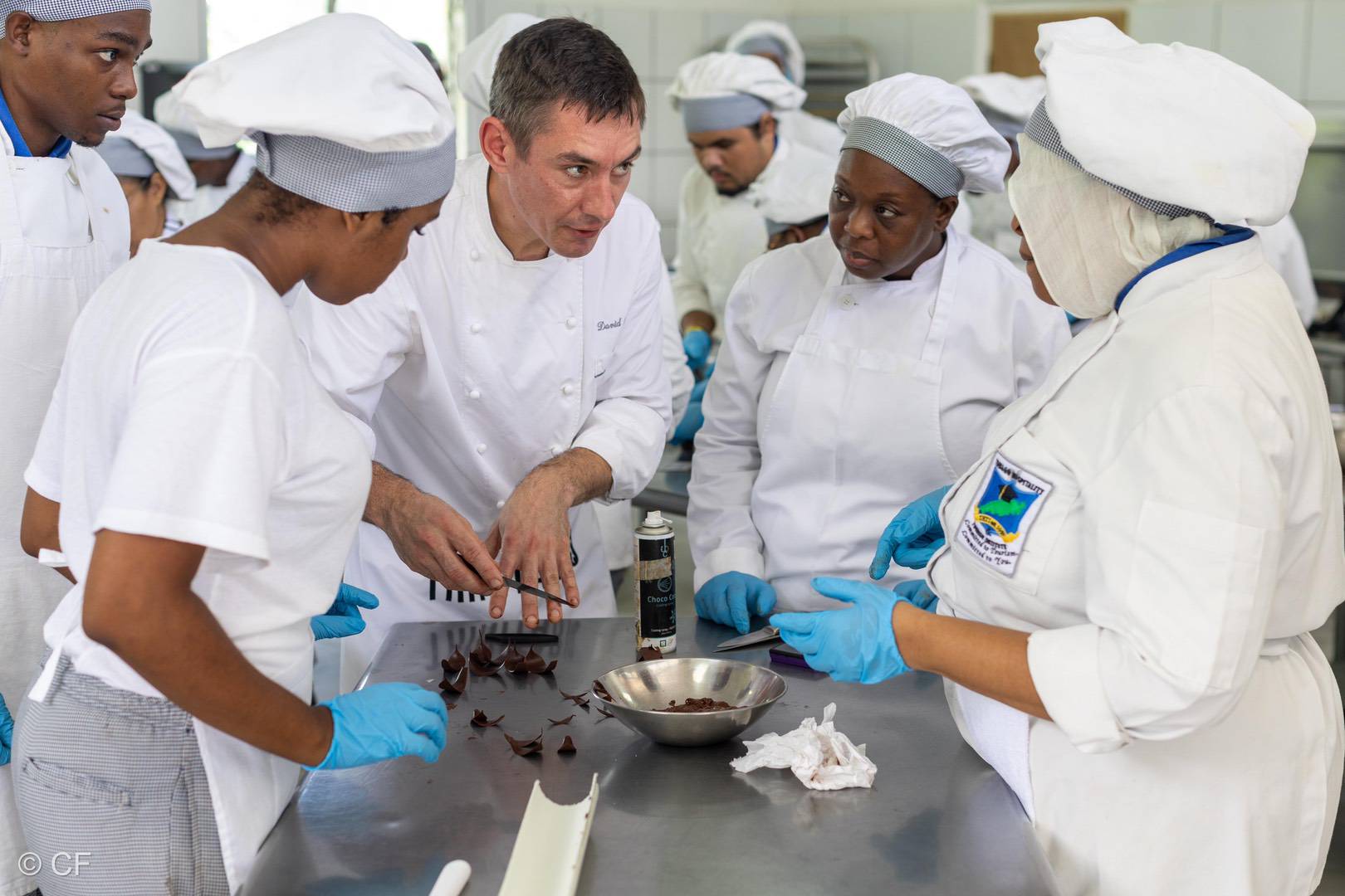A chef demonstrates a technique with fine flavour cocoa chocolate pieces to a group of students in white uniforms and blue gloves in a kitchen setting. | Trinitario
