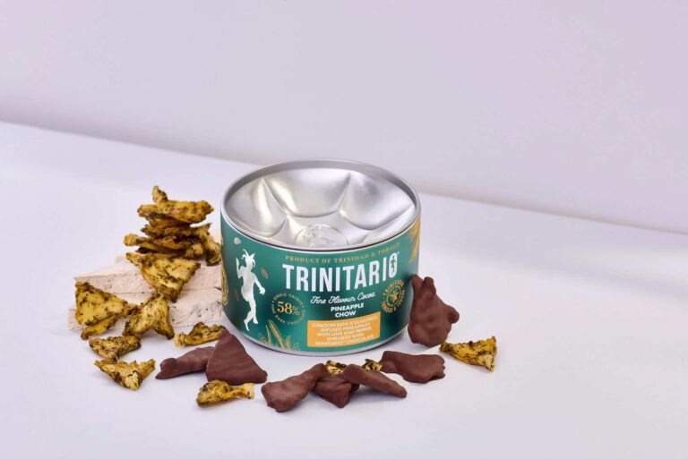 A tin labelled "Trinitario" sits on a white surface, surrounded by hand-crafted chocolate-covered pieces and irregular yellow snacks. | Trinitario