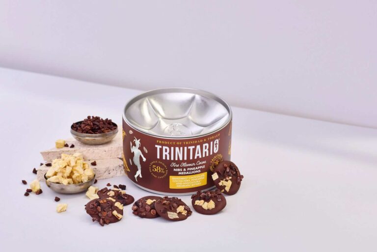 A tin of hand-crafted Trinitario chocolate with pieces of chocolate, dried fruit, and cocoa nibs scattered around on a white surface. | Trinitario