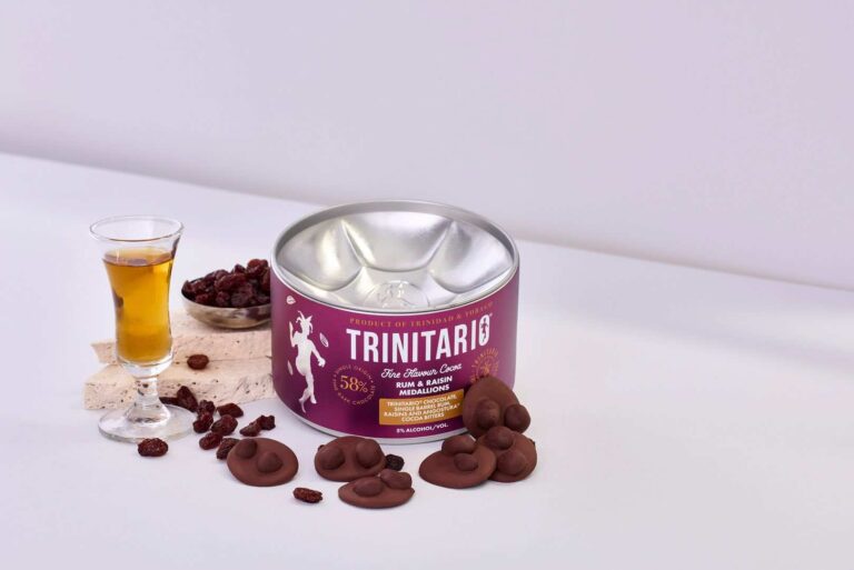 A tin of Trinitario chocolate rum and raisin medallions is displayed with chocolate pieces, dried sultanas, a glass of rum, and a textured block on a light surface, highlighting the rich taste of fine flavour cocoa. | Trinitario