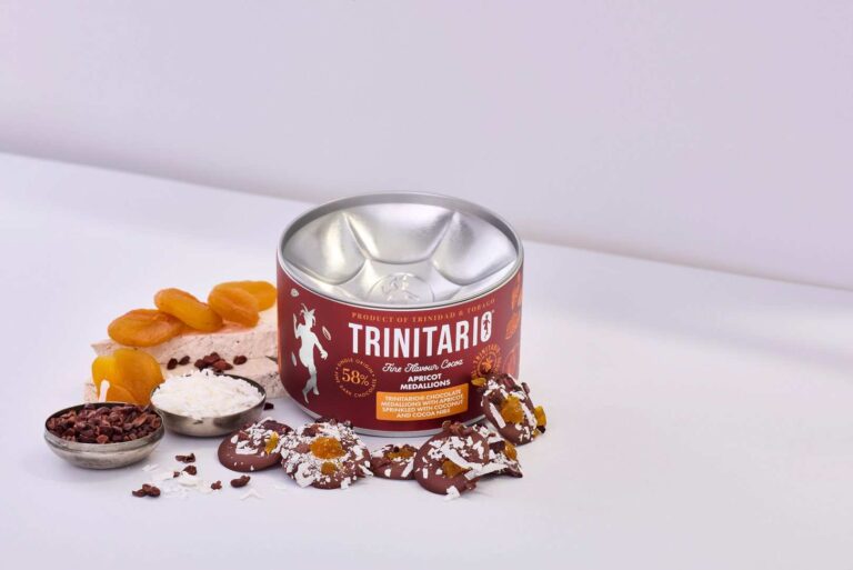 A tin of single-origin Trinitario chocolate is displayed with dried apricots, coconut, cocoa nibs, nougat, and chocolate-dipped apricot pieces topped with coconut on a white surface. | Trinitario