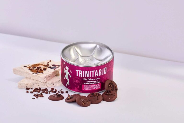 A tin of Trinitario chocolate, made from premium Caribbean cocoa, sits next to chocolate pieces, nougat, and cocoa nibs on a white surface with a pale background. | Trinitario