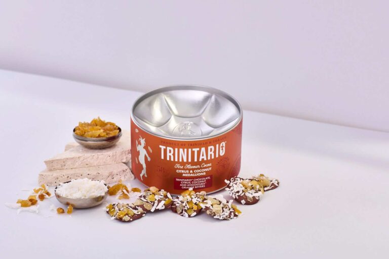 A tin of Trinitario Citrus & Coconut mineral biscuits, made with rich handcrafted chocolate and Caribbean cocoa, is displayed alongside biscuits, nougat, dried fruit, and desiccated coconut on a white surface. | Trinitario