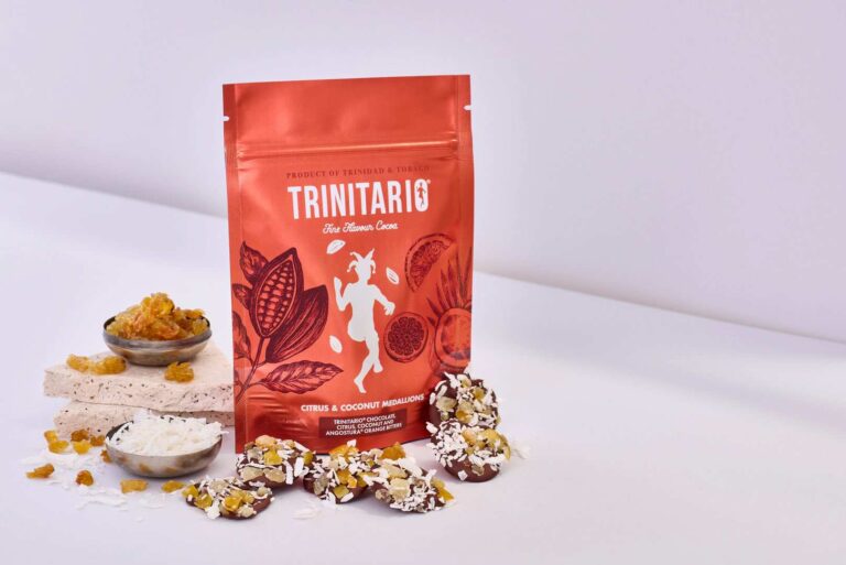 A red Trinitario Citrus & Coconut Medallion handcrafted chocolate pouch stands upright next to dried fruit, desiccated coconut, and chocolate-covered treats on a white surface. | Trinitario