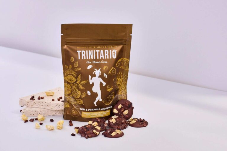 A brown parcel labelled "Trinitario" stands next to nougat pieces and hand-crafted chocolate discs topped with nuts and pineapple on a white surface. | Trinitario