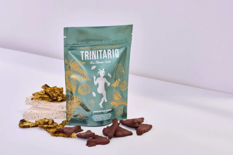 A green Trinitario Pineapple Choco snack pouch stands upright, surrounded by slices of chocolate-covered pineapple made with fine flavour cocoa and pieces of dried pineapple on a white surface. | Trinitario