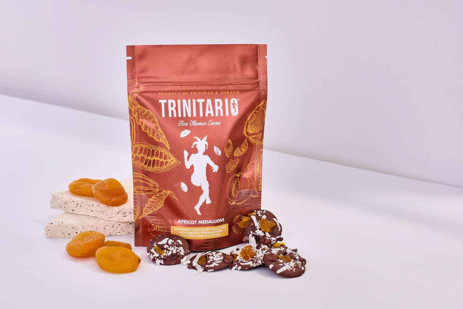 A bag of Trinitario apricot medallions, crafted with single-origin chocolate, stands upright next to dried apricots, nougat pieces, and chocolate-covered treats on a white surface. | Trinitario