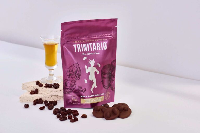 A bag of Trinitario rum and raisin medallion chocolates, made from fine flavour Caribbean cocoa, is displayed with a glass of liqueur, nougat pieces, sultanas, and chocolate discs on a white surface. | Trinitario