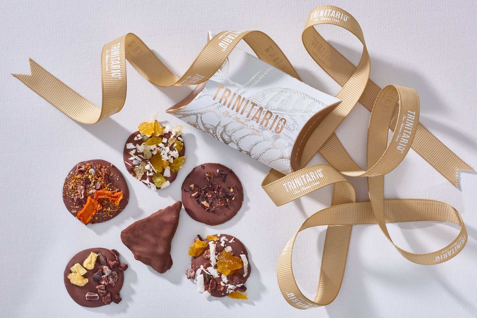 Five assorted hand-crafted chocolate-covered treats are arranged next to a decorative box and a beige ribbon printed with "TRINITARIO," showcasing the elegance of Trinitario chocolate on a light background. | Trinitario
