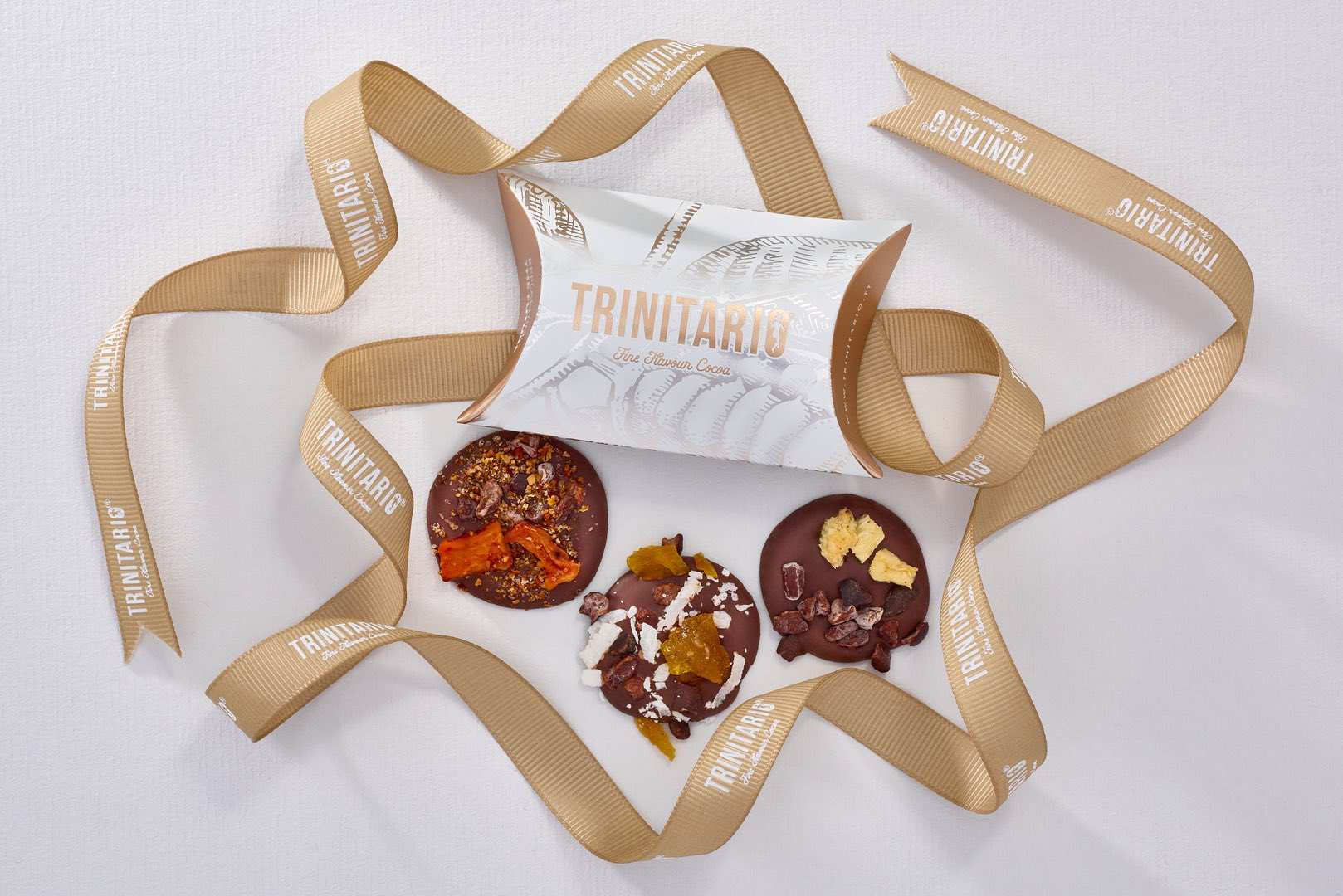 A white and gold Trinitario chocolate gift box, adorned with a branded beige ribbon and three decorated chocolate discs, showcases hand-crafted chocolate made from fine flavour cocoa on a light surface. | Trinitario
