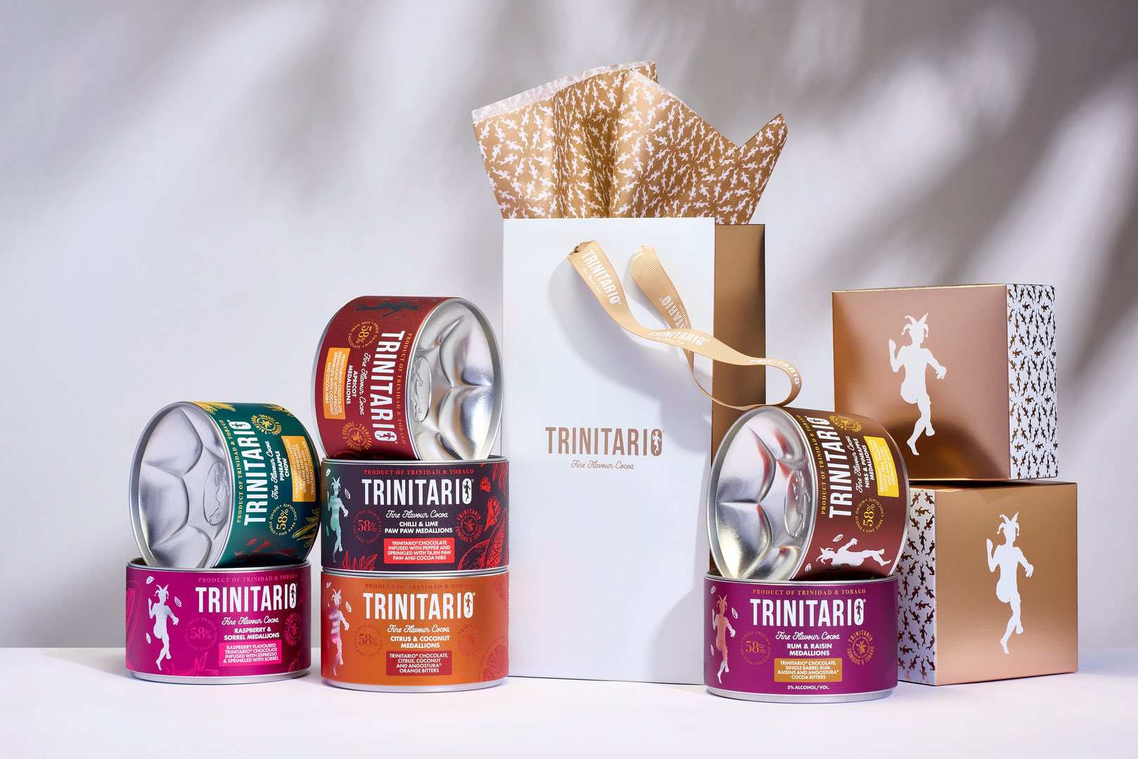 Assorted Trinitario chocolate tins, crafted from fine flavour cocoa, are arranged around a white gift bag with tissue paper and two gold gift boxes on a light surface. | Trinitario