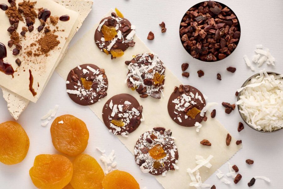 Chocolate discs made from single-origin chocolate are topped with dried apricots, desiccated coconut, and cacao nibs, then arranged on baking parchment and surrounded by more apricots, coconut, cacao nibs, and pieces of white chocolate. | Trinitario