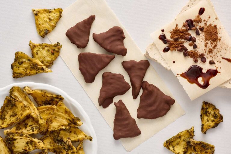 Chocolate-covered triangular snacks made with single-origin chocolate rest on baking parchment alongside dried pineapple slices and two pieces of white nougat—one topped with cocoa powder and syrup—on a white surface. | Trinitario
