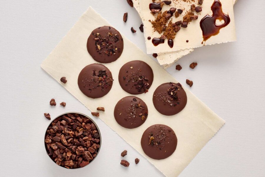 Six round dark chocolate discs made from sustainably grown Trinitario chocolate are arranged on a serviette, alongside a bowl of cocoa nibs and broken pieces of white chocolate with crumbs and chocolate drizzle. | Trinitario