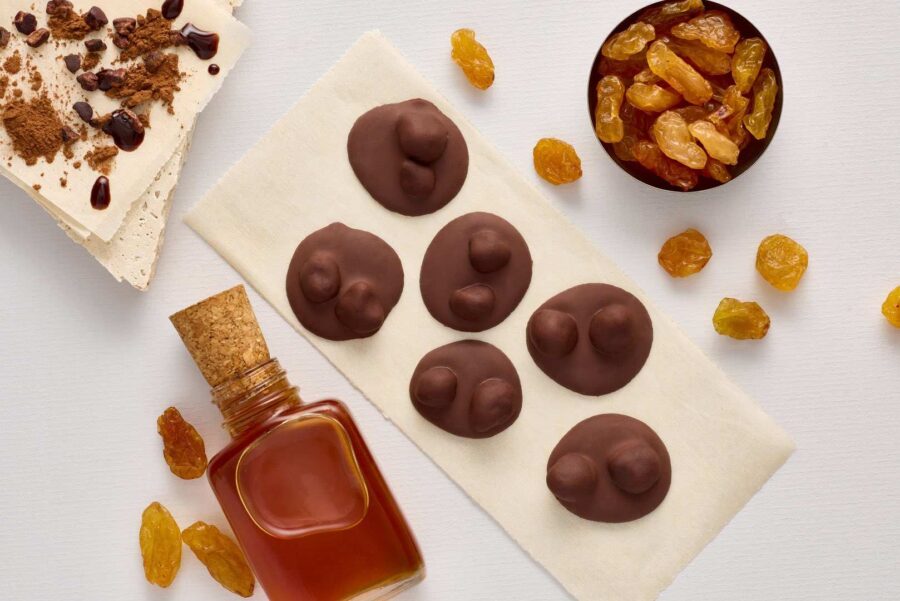 Six hand-crafted chocolate-covered clusters are arranged on a serviette, surrounded by golden sultanas, a small bottle of liquid, nougat, and a bowl of sultanas on a white surface, all featuring rich single-origin chocolate. | Trinitario