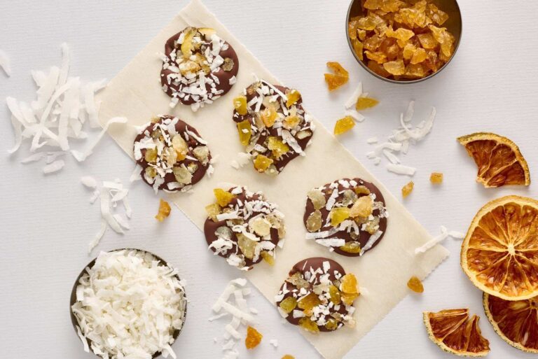 Six chocolate discs made with fine flavour cocoa are topped with shredded coconut and dried fruit, arranged on baking parchment and surrounded by bowls of coconut, dried fruit pieces, and dried orange slices. | Trinitario