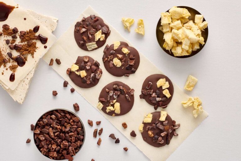 Round chocolate discs crafted from fine flavour cocoa, topped with freeze-dried pineapple and cocoa nibs, arranged on baking parchment next to bowls of the same toppings. | Trinitario