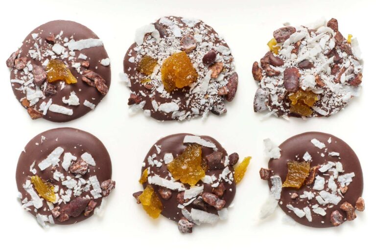 Six round pieces of hand-crafted chocolate topped with shredded coconut, cocoa nibs, and pieces of dried fruit, arranged on a white surface. | Trinitario