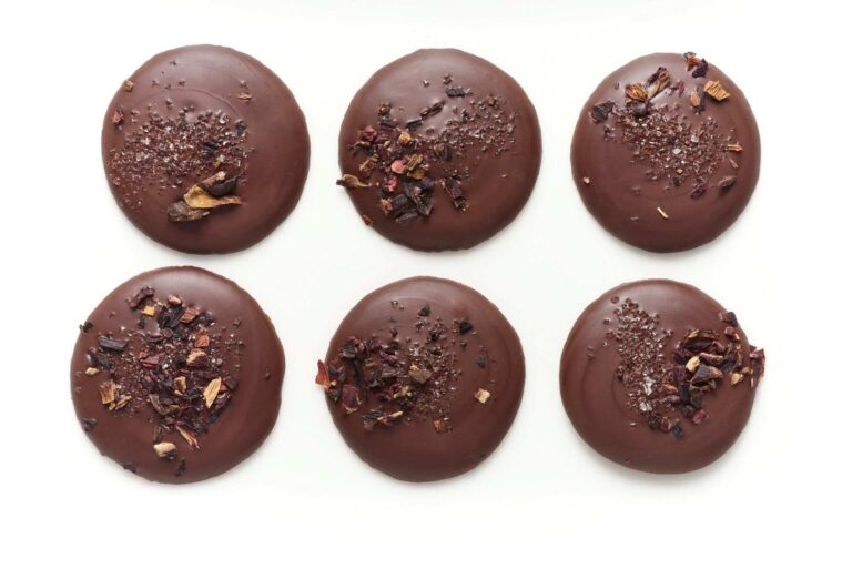 Six round biscuits, crafted with fine flavour cocoa, are coated in handcrafted chocolate and arranged in two rows of three, each topped with sea salt or dried flower petals on a white background. | Trinitario