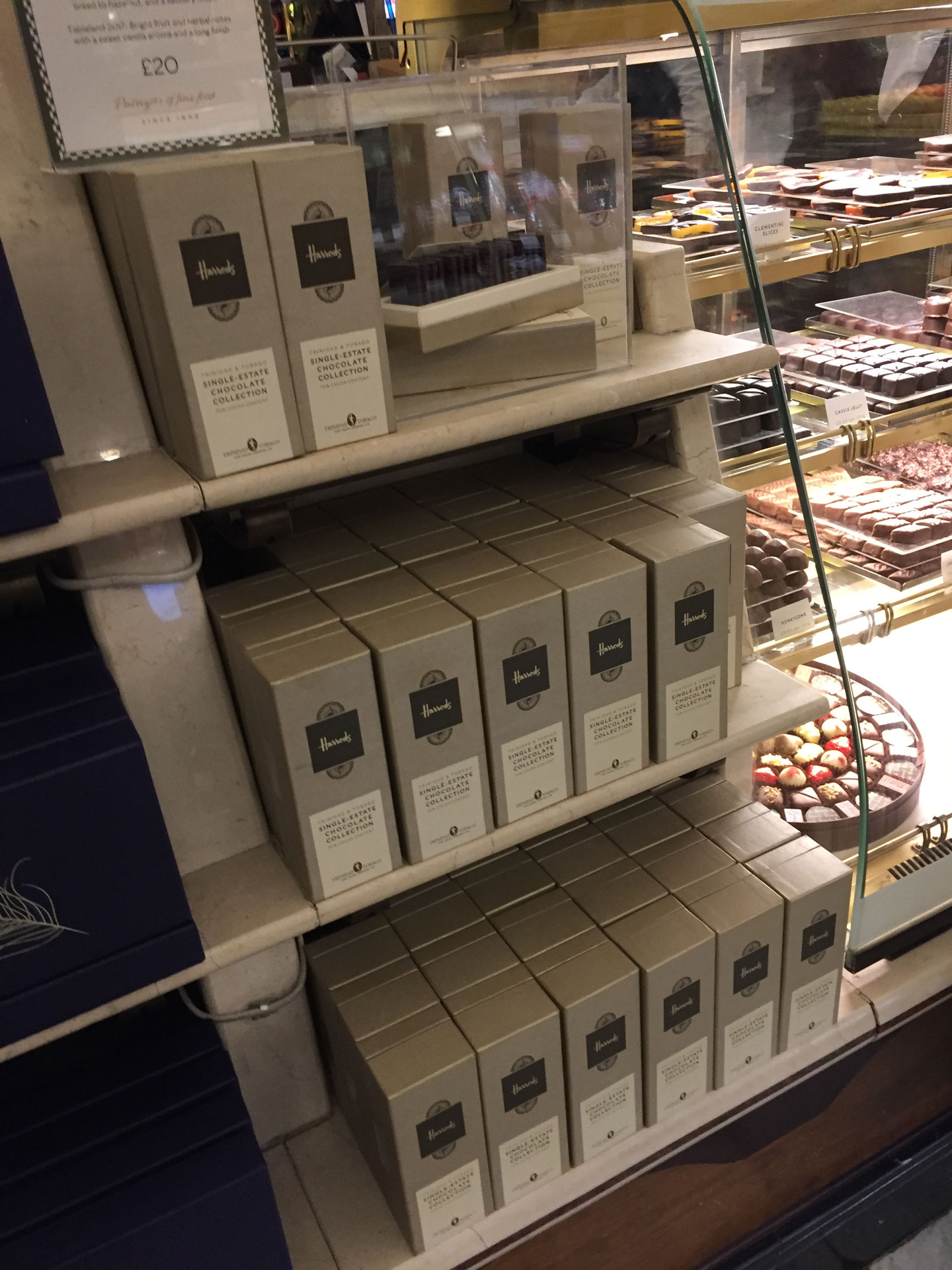 Shelves display neatly stacked beige boxes of single-origin chocolate in a shop, with various individual chocolates crafted from Caribbean cocoa visible in a glass case above. | Trinitario