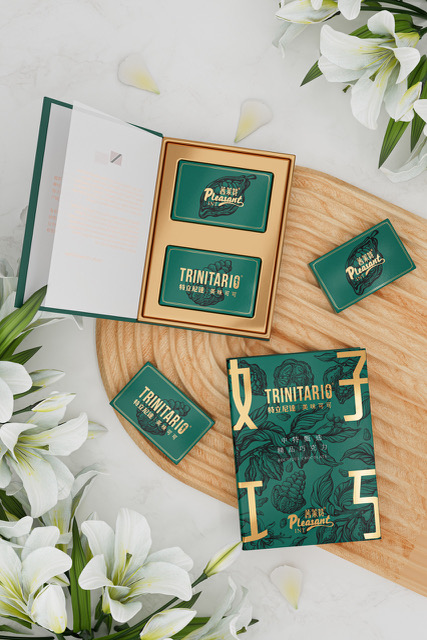 A box of Trinitario chocolate with green and gold packaging is displayed on a wooden board, surrounded by white lilies and an open booklet. | Trinitario