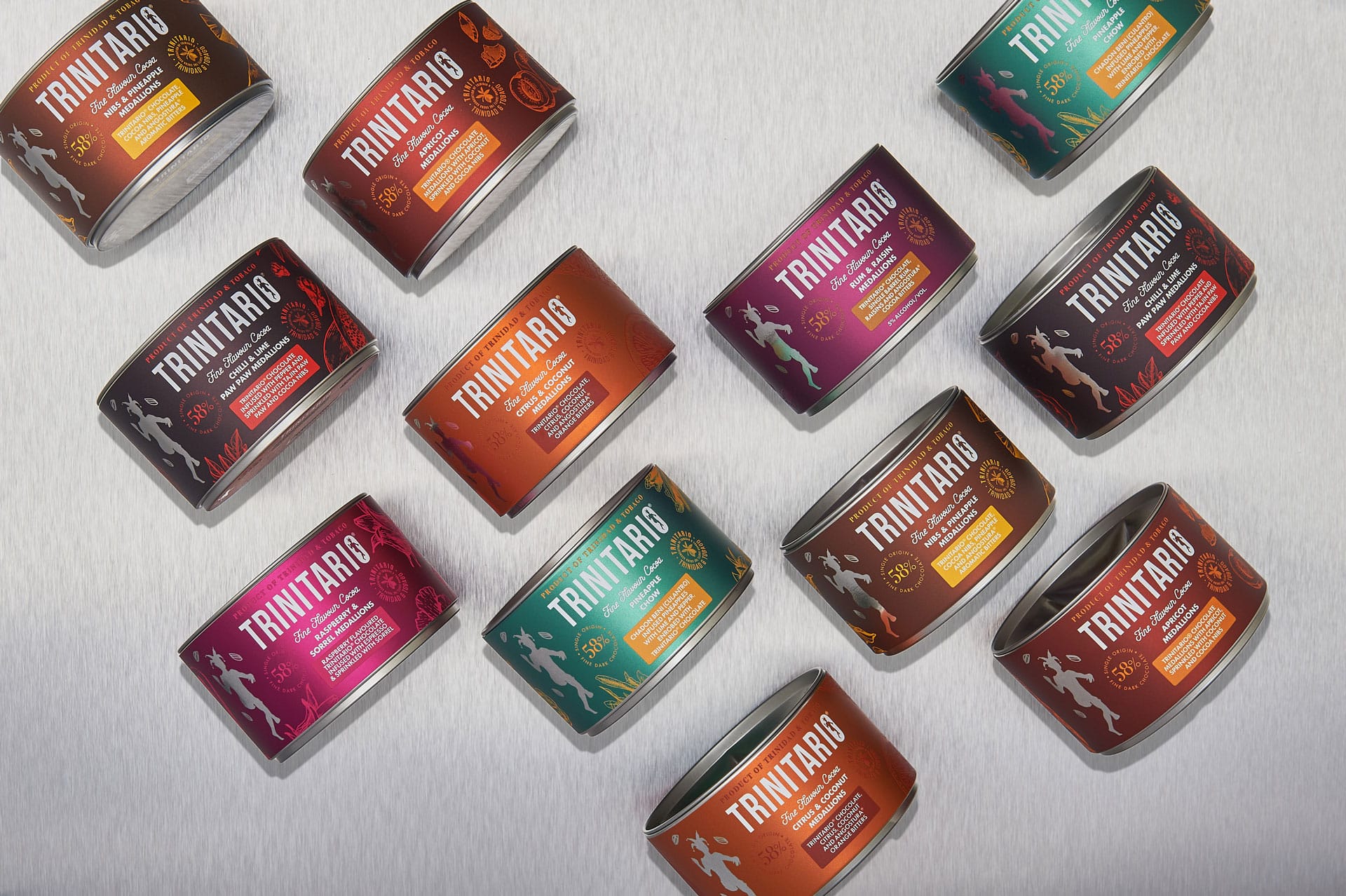 Cylindrical tins of single-origin Trinitario chocolate in various colours and flavours are arranged diagonally on a light textured surface. | Trinitario