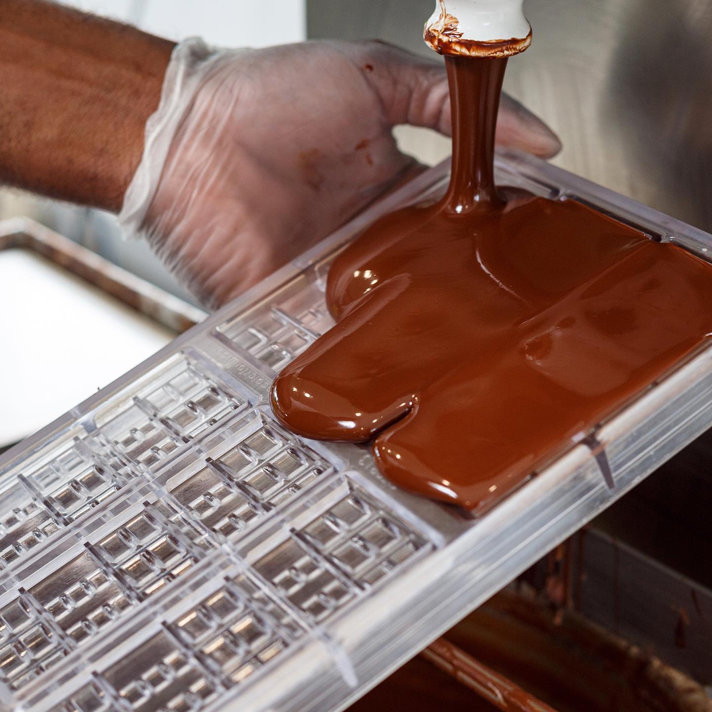 A gloved hand holds a clear plastic mould as luscious single-origin chocolate is poured into it from above, filling the compartments. | Trinitario