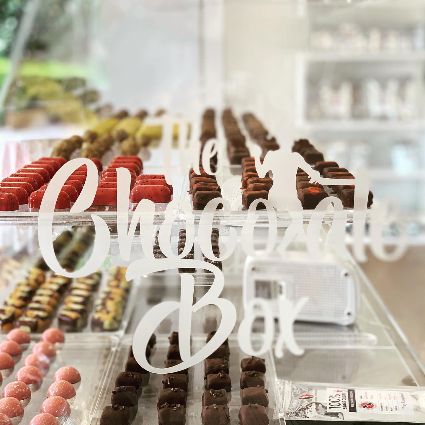 Rows of assorted chocolates made from fine flavour cocoa are displayed in a glass case inside a shop, with “The Chocolate Box” written on the glass in white script. | Trinitario