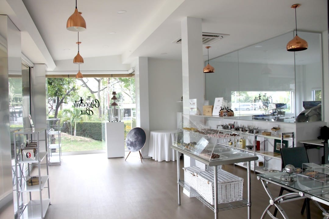 A modern, well-lit boutique with glass display cases, shelves, and pendant lights, featuring large windows and minimalist decor showcasing hand-crafted chocolate delights. | Trinitario