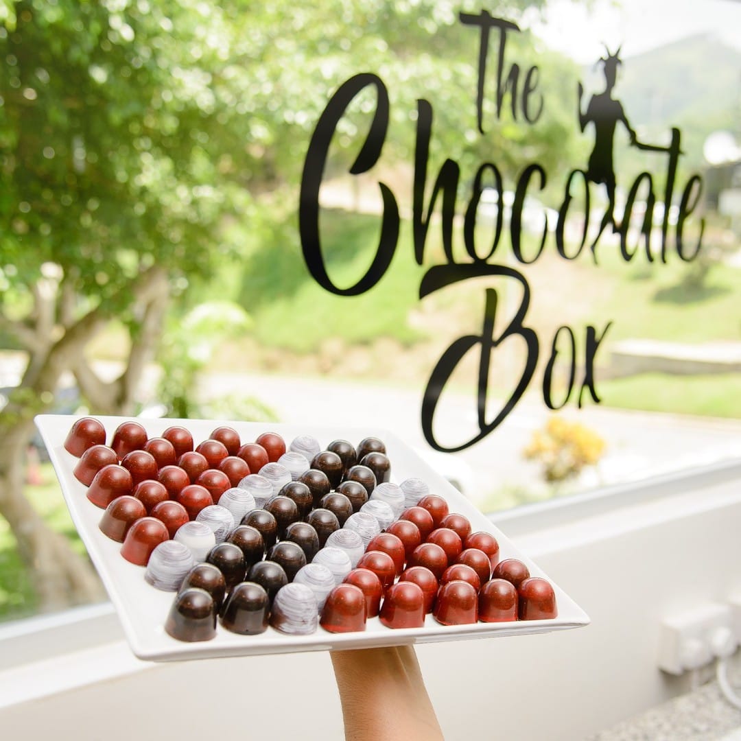 A hand holds a tray of assorted Trinitario chocolates with red, brown, and grey colours in front of a window displaying "The Chocolate Box" sign. | Trinitario
