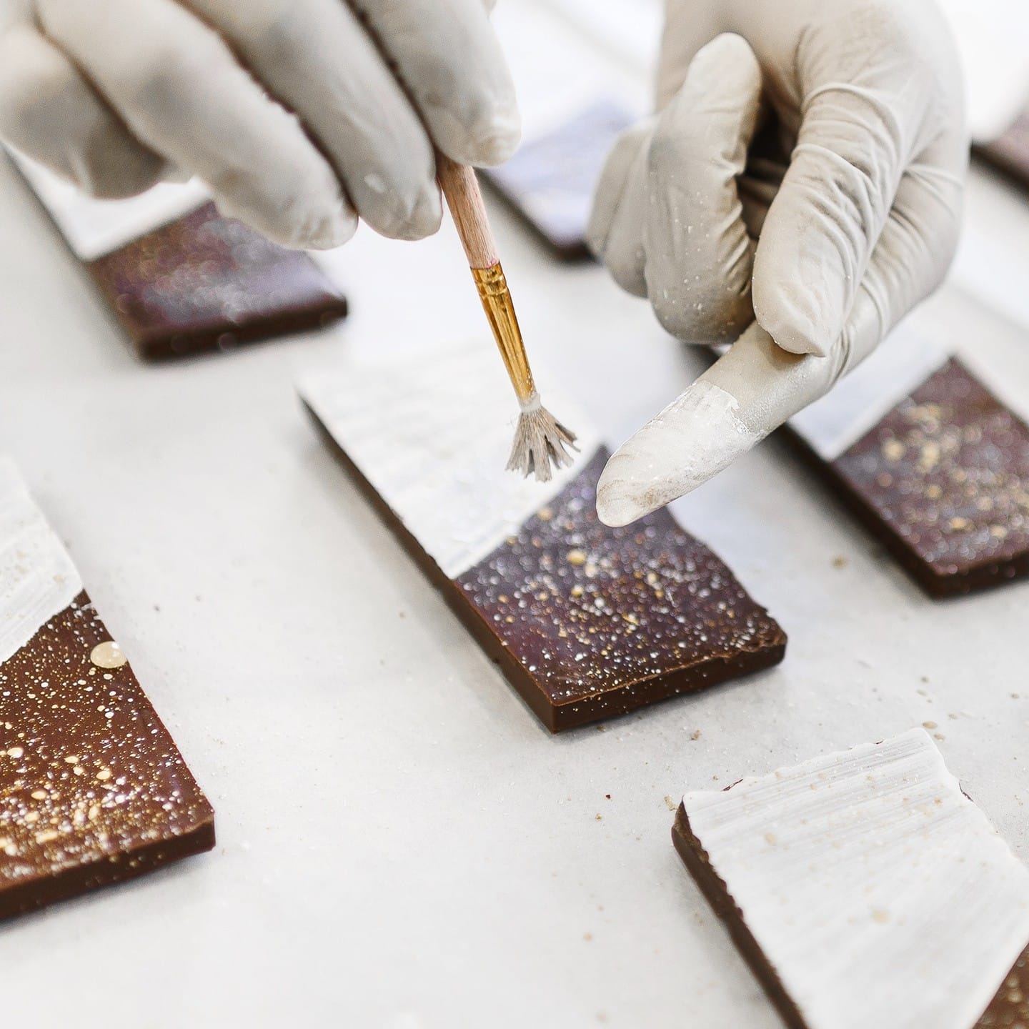 A gloved hand uses a brush to paint white edible colour onto handcrafted chocolate pieces made from fine flavour cocoa, resting on baking parchment. | Trinitario