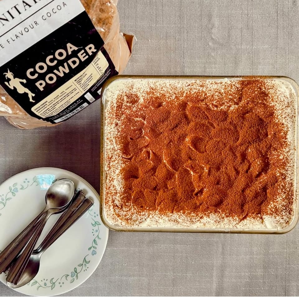 A tray of tiramisu topped with fine flavour cocoa sits next to a pack of cocoa powder, three stacked plates, and a set of four spoons on a grey surface. | Trinitario