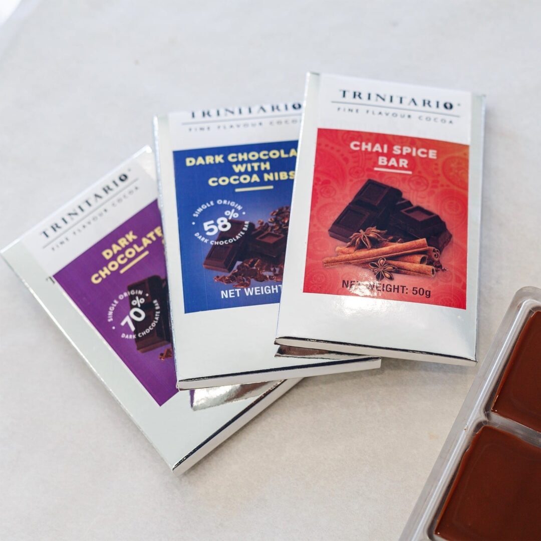 Three Trinitario chocolate bars—Dark Chocolate, Dark Chocolate with Cocoa Nibs, and Chai Spice Bar—crafted from Caribbean cocoa, are displayed next to a chocolate mould on a light surface. | Trinitario