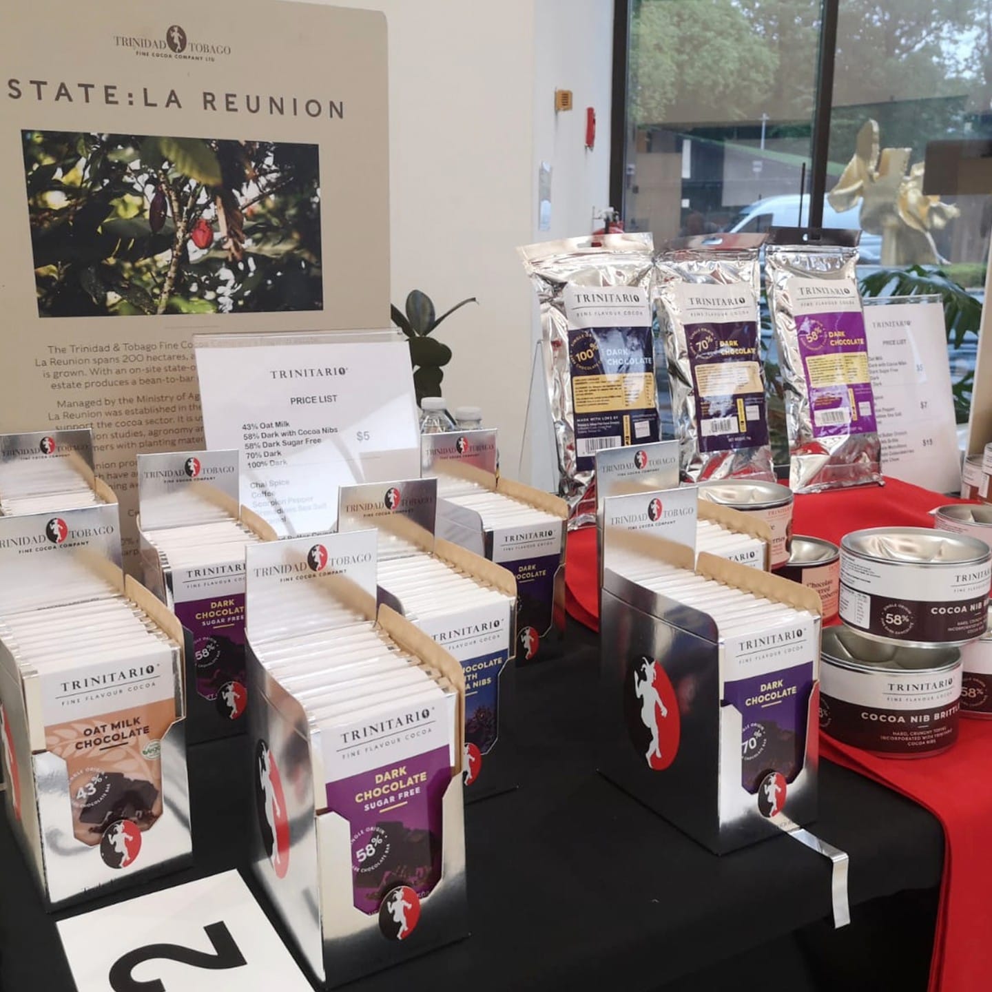 Display table featuring single-origin Trinitario chocolate products, including boxed bars, packets, tins, and informational signage highlighting the fine flavour cocoa at an event or shop. | Trinitario